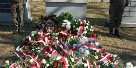 Powiększ grafikę: Na zdjęciu jest pomnik i kwiaty złożone przy nim. Wszystkie są w biało-czerwonych barwach.